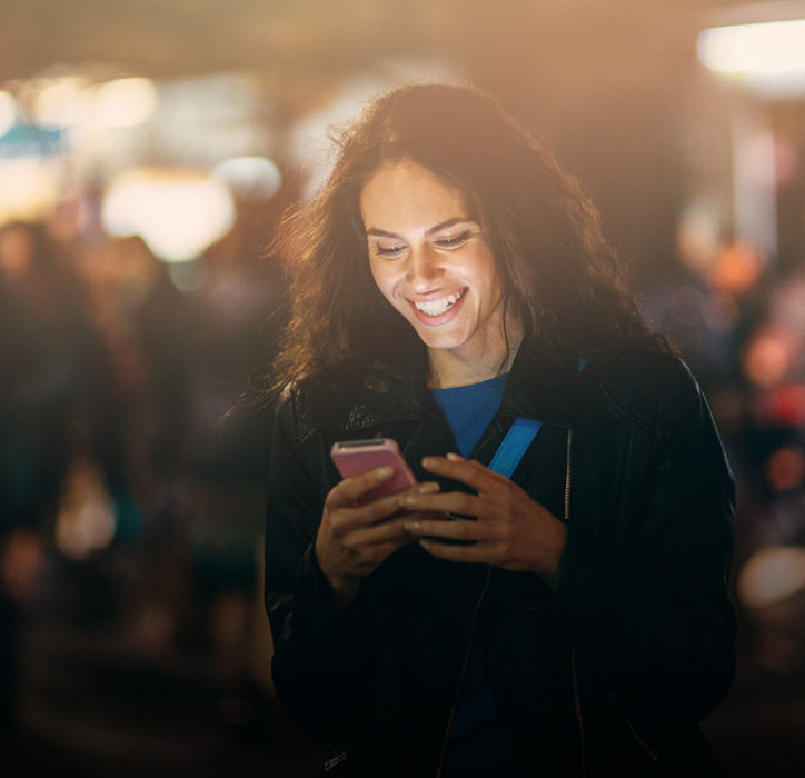 Tjej som står på gatan och ler mot sin mobiltelefon. Hon surfar fritt eftersom hon har abonnemanget Telenor Frihet Obegränsat.